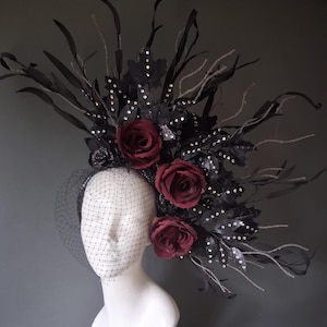 May include: A headpiece featuring three deep red roses, black leaves adorned with small clear crystals, and tall black branches. The accessory includes a black netting veil that covers the upper portion of the face, all mounted on a headband.