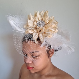 May include: A cream-colored headpiece featuring a large fabric flower with a rhinestone center, white feathers, and a delicate net veil. This elegant accessory is suitable for special occasions.