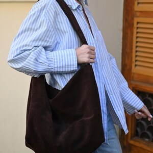 Może przedstawiać: Ciemnobrązowa torba typu hobo z zamszu z szerokim paskiem na ramię, noszona na koszuli w jasnoniebieskie i białe pionowe paski oraz jasnoniebieskich dżinsach. Torba ma miękki, luźny fason oraz matowe, teksturowane wykończenie powierzchni.