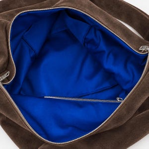 Może przedstawiać: Brązowa torba typu hobo z zamszu z wyści&oacute;łką z tkaniny w kolorze żywego kr&oacute;lewskiego błękitu. Torba posiada srebrny metalowy zamek błyskawiczny przy g&oacute;rnym otworze oraz dodatkową wewnętrzną kieszeń na zamek, ukazującą przestronną komorę.