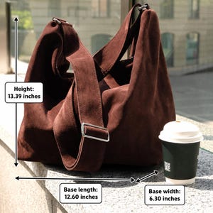 Może przedstawiać: Ciemnobrązowa torba typu hobo z zamszu z szerokim paskiem na ramię i srebrnymi okuciami. Torba ma 34 cm wysokości, 32 cm długości podstawy i 16 cm szerokości podstawy. Obok znajduje się ciemnozielony jednorazowy kubek na kawę.