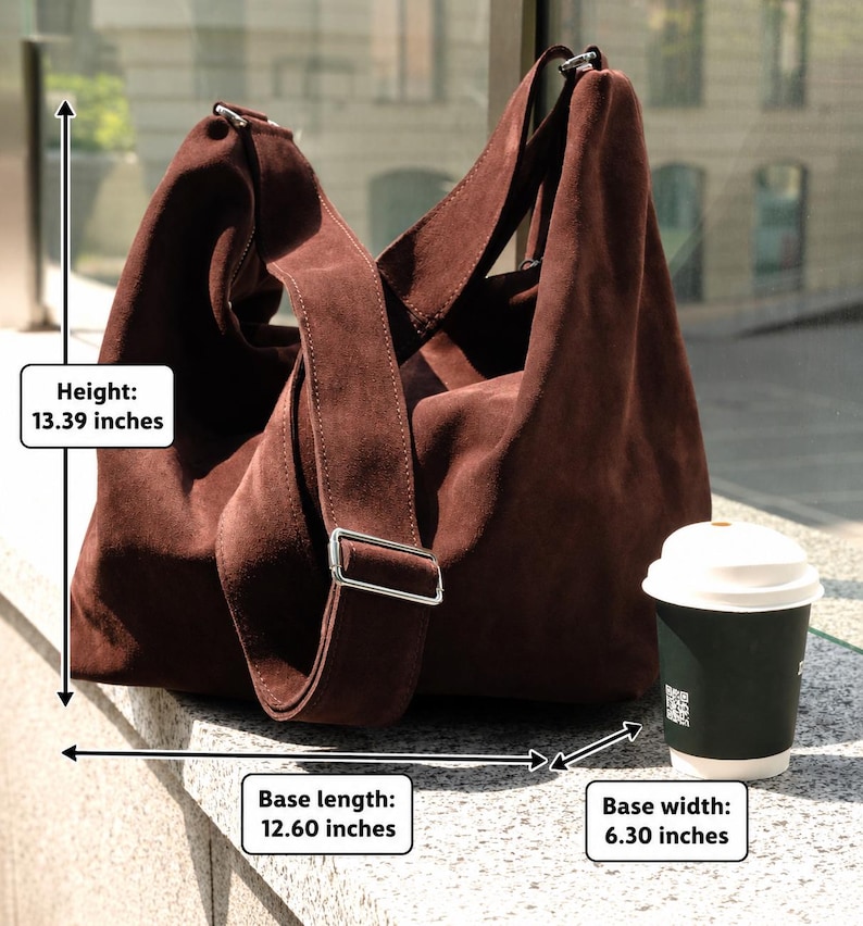 Może przedstawiać: Ciemnobrązowa torba typu hobo z zamszu z szerokim paskiem na ramię i srebrnymi okuciami. Torba ma 34 cm wysokości, 32 cm długości podstawy i 16 cm szerokości podstawy. Obok znajduje się ciemnozielony jednorazowy kubek na kawę.