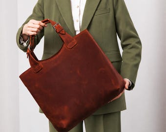 May include: A large, square-shaped tote bag in a deep reddish-brown color. The bag features two sturdy handles attached with silver-toned metal buckles, allowing for adjustable length. The surface has a matte, smooth texture with subtle variations.