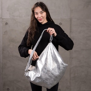 Może przedstawiać: Metaliczna srebrna torba typu hobo o teksturowanym, pogniecionym wykończeniu. Torba posiada pojedynczy szeroki pasek na ramię oraz luźną, trójkątną sylwetkę. Materiał ma odblaskową, błyszczącą powierzchnię, która odbija światło.