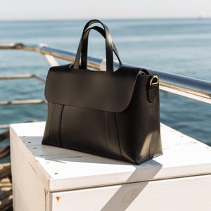 以下が含まれることがあります： フラップ開閉と2つのトップハンドルが付いた黒い革のハンドバッグ。バッグは、側面に金色の金属アクセントが付いた構造的なデザインです。背景には海と白い表面の景色があります。