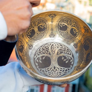 May include: A golden singing bowl with intricate tree of life designs etched into its surface. The bowl's rim and interior feature detailed patterns, creating a visually rich and spiritual aesthetic. The bowl is held by a person.