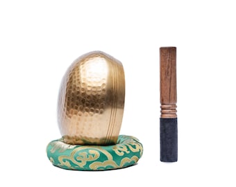 May include: A gold singing bowl with a hammered surface sits on a green cushion with a gold pattern. A wooden mallet with a black rubber tip is included.