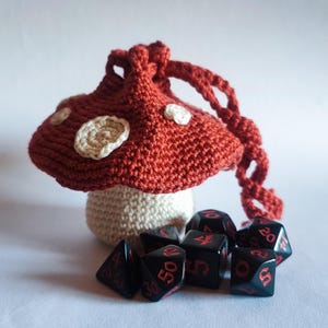 May include: A handmade crochet mushroom dice bag in red with white spots, and a cream-colored stem. Black dice with red numbers are scattered in front of the bag. The bag has a drawstring closure. The theme is fantasy and gaming.