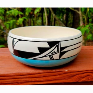 May include: A round ceramic bowl featuring a cream-colored interior and a white exterior decorated with black geometric line art. The base of the bowl is painted in a solid bright turquoise blue, creating a distinct contrast with the upper section.
