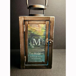 May include: A rectangular lantern with a distressed bronze finish and a top handle. The glass panel features an etched letter M inside a circular wreath, followed by the text The Malkins, JoAnn & David, and the date 6-22-2008 in white lettering.