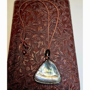 May include: A triangular pendant necklace featuring an iridescent abalone shell inlay set in a dark metal frame. The pendant hangs from a thin brown cord with adjustable sliding knots and two small round brown wooden beads for length control.
