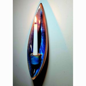 May include: A wall-mounted ceramic candle holder with an elongated, leaf-like shape. The glaze features deep blue and brown tones with a glossy finish. A single white taper candle is inserted into the central cup, emitting a warm flame.