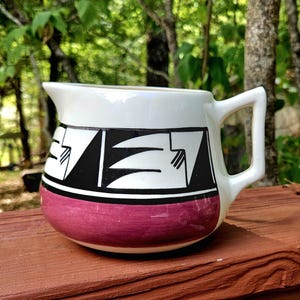 May include: A ceramic creamer pitcher featuring a cream-colored upper body with a black geometric pattern and a solid magenta band around the base. The pitcher has a wide pouring spout and a sturdy angular handle on the right side.