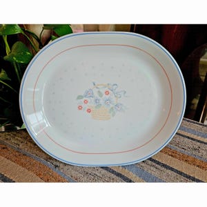 May include: An oval white ceramic serving platter featuring a central illustration of a woven basket filled with blue and red flowers tied with a light blue ribbon. The rim is decorated with thin concentric blue and pink circular lines.