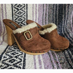 May include: Brown suede clogs featuring a high wooden heel, a brass-toned buckle strap across the top, and a cream-colored faux fur trim around the collar. The base of the shoe is accented with small metal studs along the wooden platform.