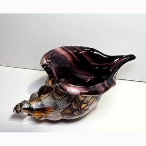 May include: A decorative glass sculpture shaped like a conch shell, featuring swirling patterns of deep purple, amber, and clear glass. The object has a smooth, glossy finish with a wide, open aperture and a tapered, pointed end.