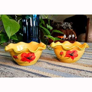 May include: Two small, yellow ceramic bowls with ruffled, wavy edges. Each bowl features a hand-painted red poppy flower design with green stems and dark centers. The yellow glaze has a subtle speckled texture throughout the exterior surface.