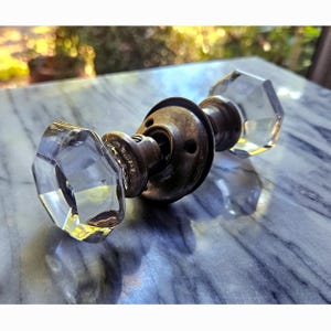 May include: A vintage door knob set featuring two clear, multifaceted glass knobs attached to a central, tarnished brass-toned metal spindle and circular mounting plate. The glass components have a distinct octagonal shape with reflective surfaces.