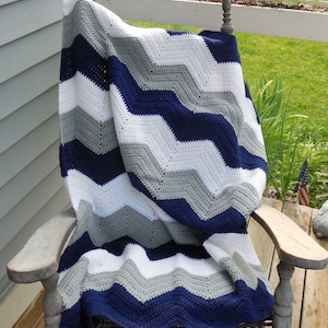 May include: A handmade crochet blanket draped over a wooden rocking chair. The blanket features a chevron pattern in navy blue, white, and gray. The blanket is soft and cozy, perfect for snuggling up on a chilly day.