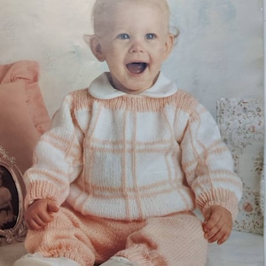 Könnte beinhalten: Ein Baby trägt einen gestrickten Pullover mit rosa und weißen Karos und eine passende Hose. Das Baby sitzt auf einer weißen Oberfläche und lächelt.