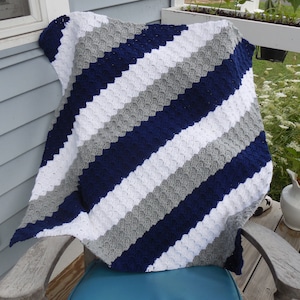 Peut inclure: Une couverture pour bébé en crochet avec un motif de rayures diagonales en blanc, gris et bleu marine.