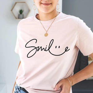 May include: A pink t-shirt with a black cursive design that says "Smile..." with a smiley face incorporated into the cursive.