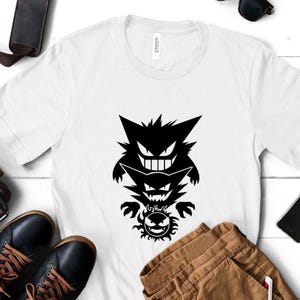 May include: A white short-sleeved t-shirt featuring a black graphic print of three stacked creatures. The top figure is a large, grinning, pointed-eared character, the middle is a smaller version, and the bottom is a circular, spiky-haired face.