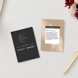 May include: Two cards with text on a white surface. One card is black with gold text that reads "Cosmic Navigator" and "The 10 Universal Laws CHEAT SHEET". The other card is tan with text about the Law of Attraction. A gold pen and a pink vase with red flowers are also present.