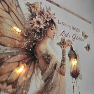 May include: Wall art depicting a fairy with illuminated wings and a lantern. The fairy has floral hair and tattoos. The text "Shine Bright Like Glitter" is visible. The artwork has a warm, inviting colour palette.
