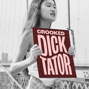 May include: A person holding a sign that says "Crooked Dicktator" in pink letters on a maroon background. The sign is surrounded by a border of small white stars.