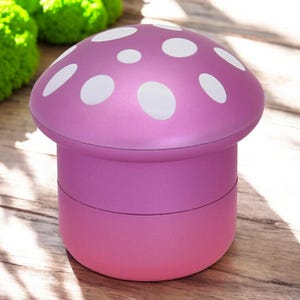 Pink mushroom-shaped grinder with white polka dots, shown closed on a wooden surface with a natural light background – a cute and funny 4-piece spice grinder for herbs, ideal as a gift or kitchen accessory.