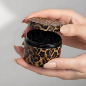 Puede incluir: Un molinillo de hierbas cilíndrico con un estampado de leopardo en tonos marrones y negros en el exterior. El dispositivo consta de una tapa extraíble y un cuerpo principal con dientes interiores negros visibles para moler hierbas.
