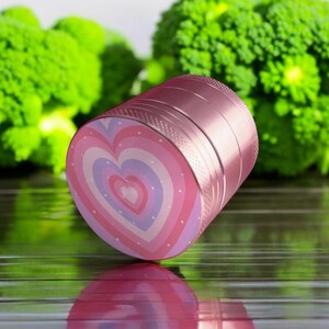 Compact pink and purple-toned heart-themed herb grinder lying horizontally, perfect for tobacco, stash jar accessory, or gift for her.