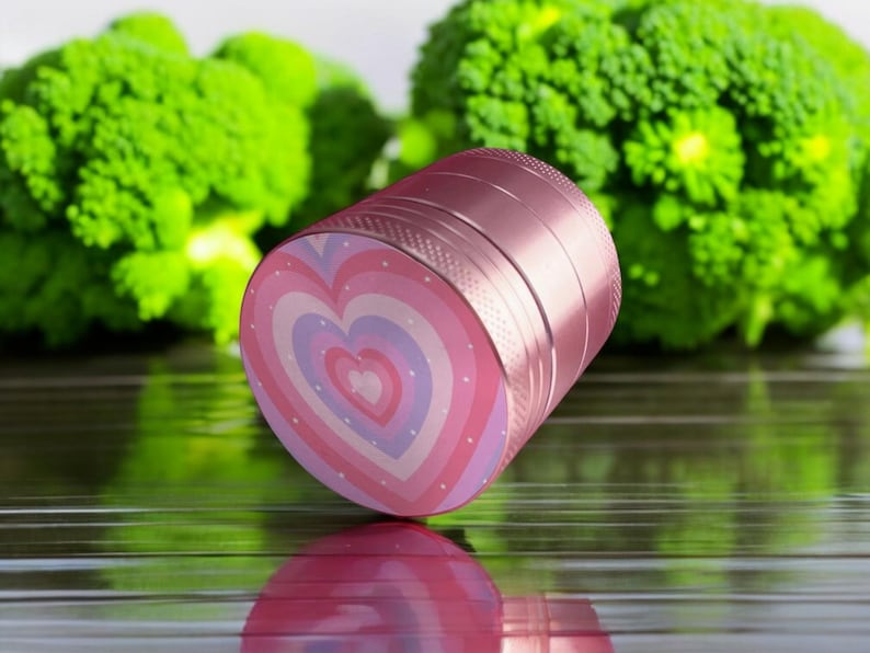 Compact pink and purple-toned heart-themed herb grinder lying horizontally, perfect for tobacco, stash jar accessory, or gift for her.