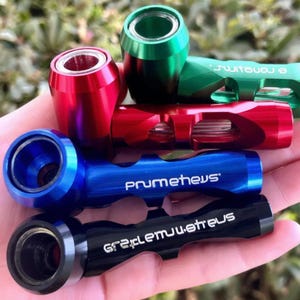 Op de afbeelding: Een assortiment kleurrijke metalen pijpen in rood, groen, blauw en zwart. Op de blauwe pijp staat het woord "prumetheus" gedrukt. Op de zwarte pijp staat het woord "gretclemu.etreu" gedrukt. De pijpen zijn cilindrisch met een cirkelvormige opening.