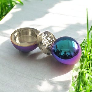 Peut inclure: Un moulin à herbe iridescent métallique en forme de sphère, avec un couvercle amovible et un mécanisme de broyage. Le moulin est ouvert, révélant le mécanisme de broyage.