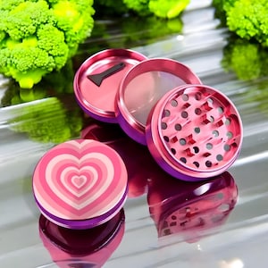 May include: A multi-piece metal herb grinder with a vibrant pink and purple gradient finish. The top lid features a concentric heart pattern in shades of pink and white. The interior components show sharp teeth for grinding and a small black scraper.