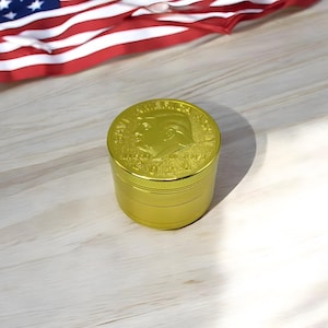 Puede incluir: Un molinillo de hierbas cilíndrico de metal dorado con un perfil en relieve de Donald Trump en la tapa, con el texto "SAVE AMERICA AGAIN" arqueado sobre el retrato y "MADE IN USA" inscrito debajo del retrato en la superficie superior.