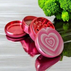 Open 4-piece pink grinder with heart design, sharp metal teeth and mesh screen, great for grinding herbs and spices.