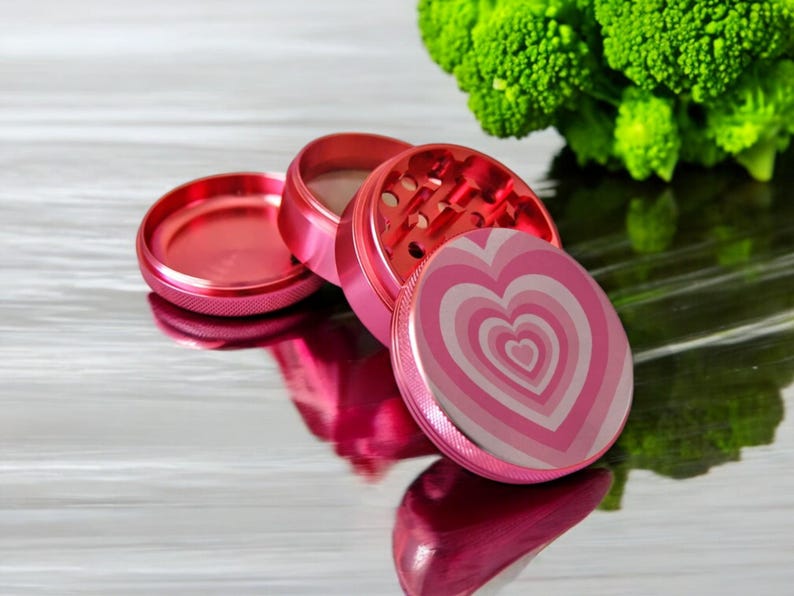 Open 4-piece pink grinder with heart design, sharp metal teeth and mesh screen, great for grinding herbs and spices.