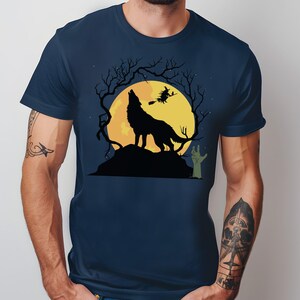May include: Navy blue t-shirt featuring a Halloween design. A black silhouette of a howling wolf is centered on a large yellow moon, with a witch flying on a broomstick. Bare tree branches and a green zombie hand complete the spooky scene.