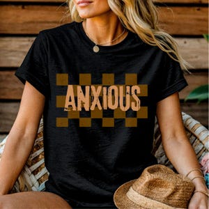 May include: Black t-shirt with the word "ANXIOUS" in a distressed, tan font, set against a brown and tan checkered background. The shirt has short sleeves and a crew neck.