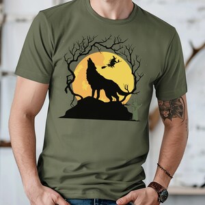 May include: Olive green t-shirt featuring a Halloween-themed graphic. The design includes a howling wolf silhouette on a hill, a full yellow moon, bare tree branches, and a witch flying on a broomstick. The shirt is made of a soft material.