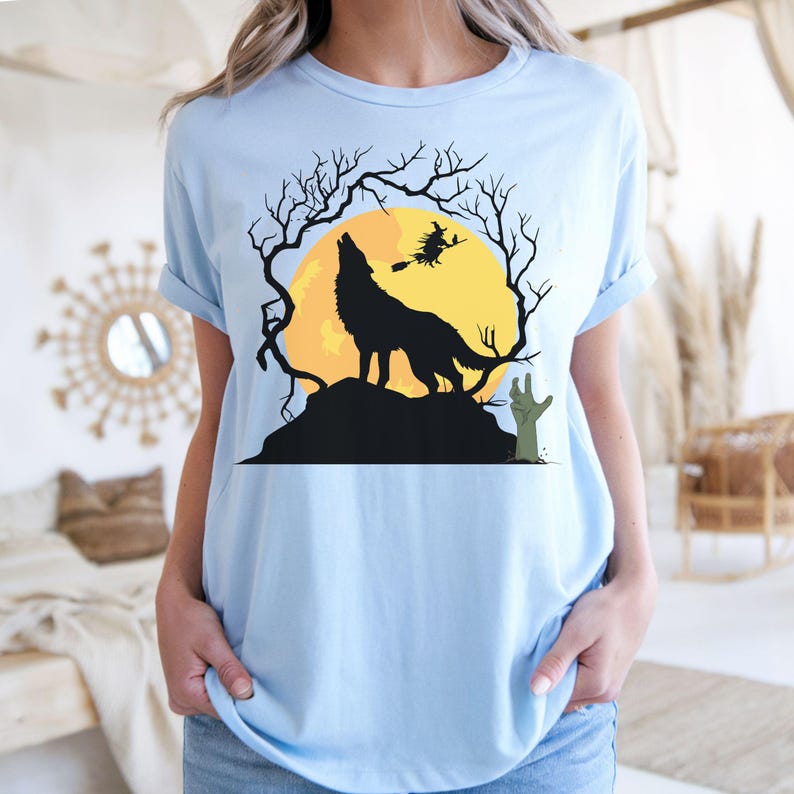 May include: Light blue t-shirt featuring a Halloween-themed graphic. The design includes a howling wolf silhouette against a yellow moon, with a witch flying nearby. Bare tree branches and a zombie hand complete the spooky scene.