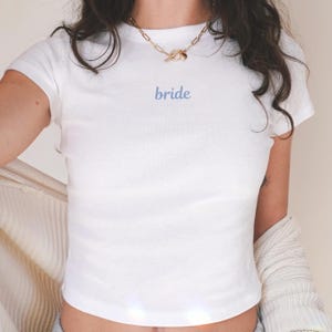 Może przedstawiać: Biały t-shirt z krótkim rękawem z wyhaftowanym słowem "bride" w jasnoniebieskim kolorze. Koszulka ma dopasowany krój. Widoczny jest złoty łańcuszek. Ogólny motyw to odzież ślubna.