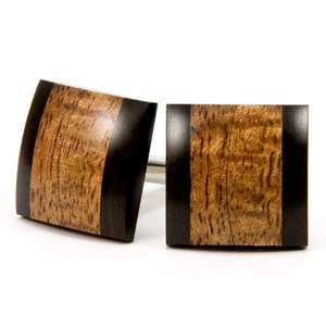 May include: A pair of square wooden cufflinks with a black inlay. The cufflinks have a wood grain pattern and a dark brown color.