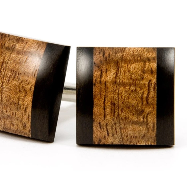 May include: A pair of square cufflinks made of wood with a black border. The wood has a brown and gold color with a natural grain pattern.