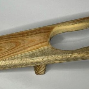 May include: A wooden boot jack with a natural wood finish. The jack has a light brown color with darker brown streaks throughout the wood.