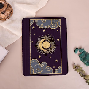 May include: A rectangular dark purple tablet case featuring a gold foil design of a central sun and crescent moon surrounded by stars and stylized grey clouds. The illustration is framed by a thin gold border on the front cover.