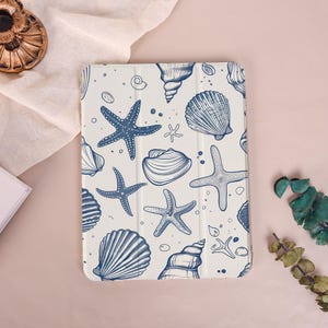 Könnte beinhalten: Weiße Tablet-Hülle mit blauem Muschel- und Seesternmuster. Das Design zeigt verschiedene Muscheln und Seesterne in einem detaillierten, handgezeichneten Stil. Die Hülle ist ein schützendes Zubehör für ein Tablet.